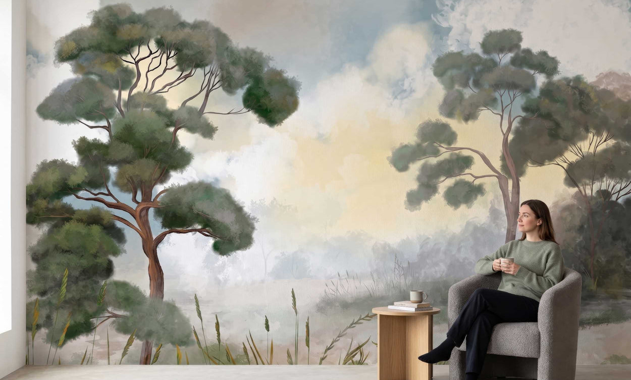 Woman sitting in a chair in front of a wall mural of a scenic landscape with trees and clouds.