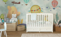 White crib placed against a sky-themed cartoon wall mural with colorful bunting.