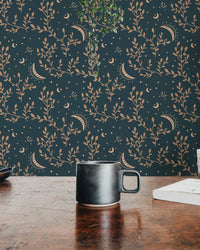 Dark teal wallpaper with gold celestial moon and floral patterns behind a coffee mug.