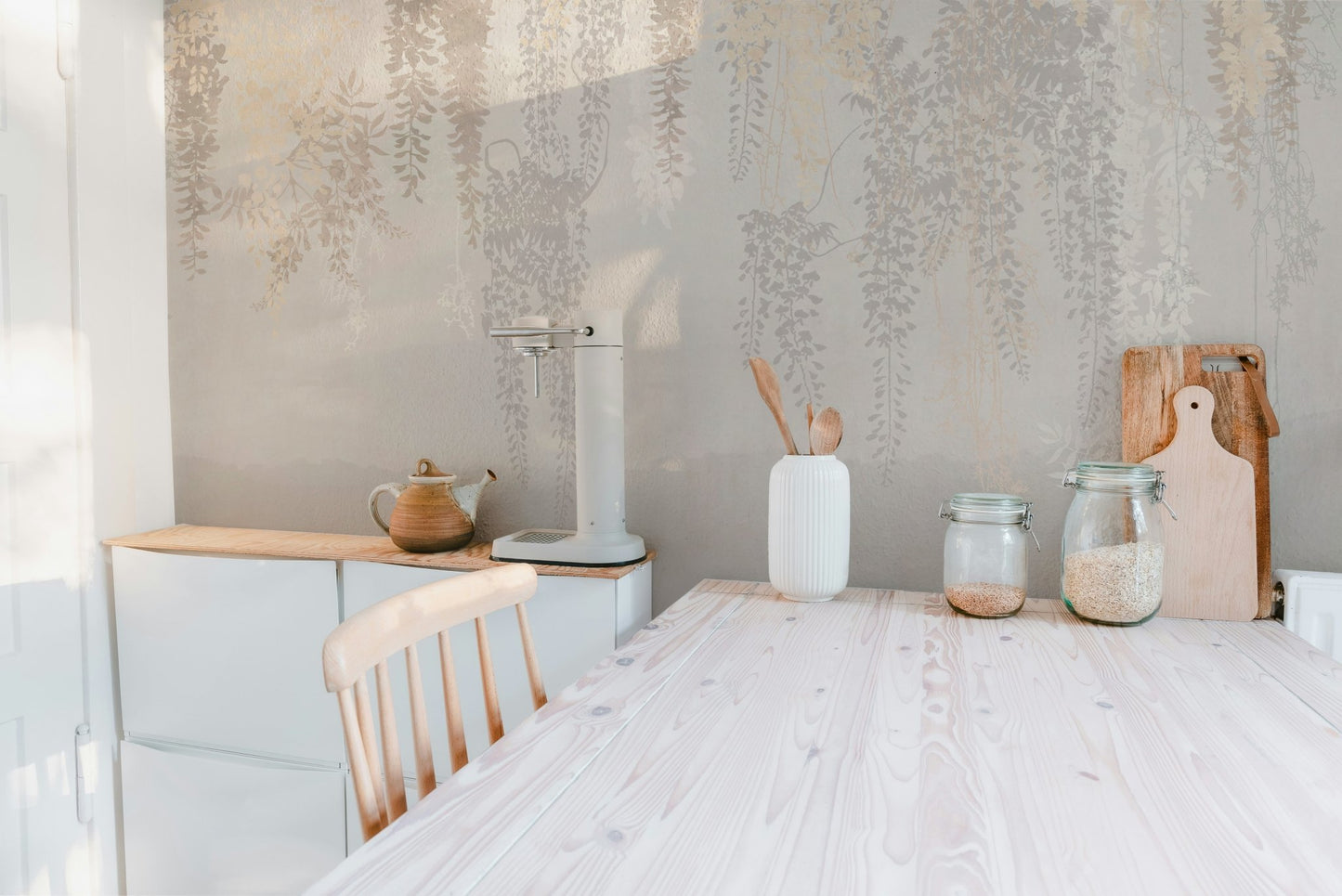 Neutral leaves pattern wall mural behind a wooden dining table and kitchen jars.