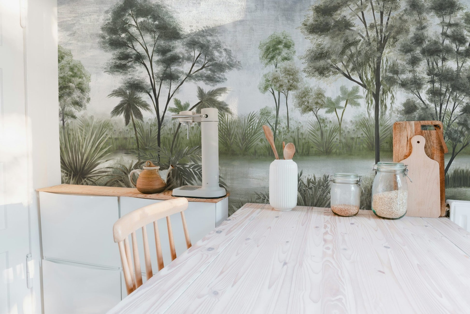 Forest landscape wall mural behind a light wood dining table in kitchen.