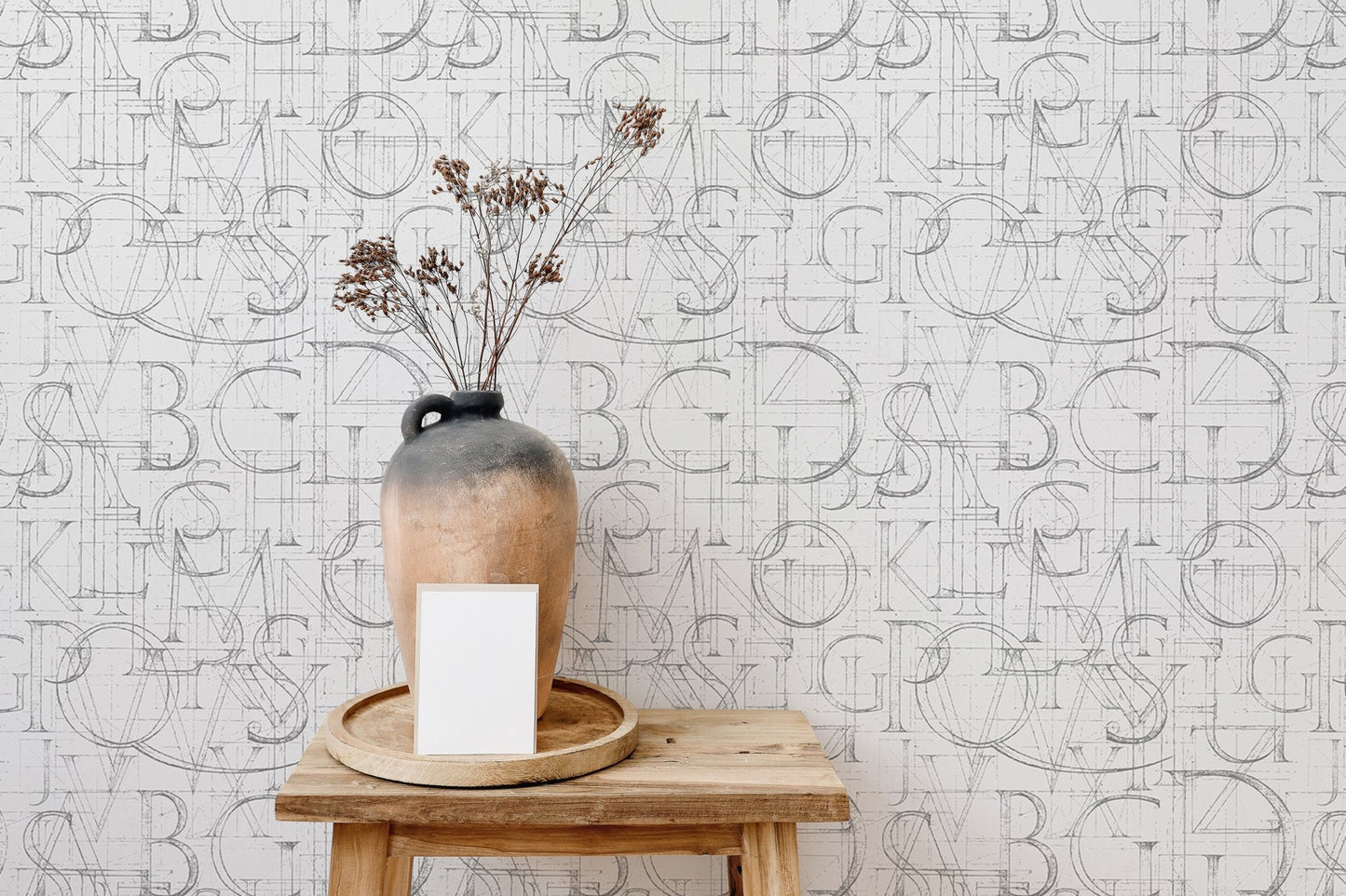 Minimalist alphabet wallpaper behind a small wooden stool and dried flora.