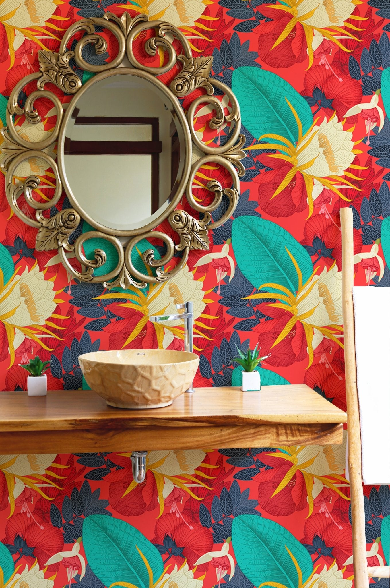 Red tropical wallpaper behind a round gold mirror and wooden shelf in bathroom.