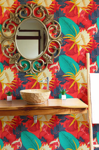 Red tropical wallpaper behind a round gold mirror and wooden shelf in bathroom.