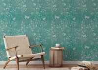 Turquoise wallpaper with botanical prints in a sunlit reading area with a side table.
