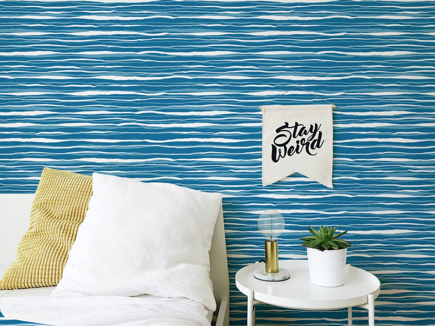 Bedroom wall with blue ocean waves wallpaper behind a white pillow and side table.