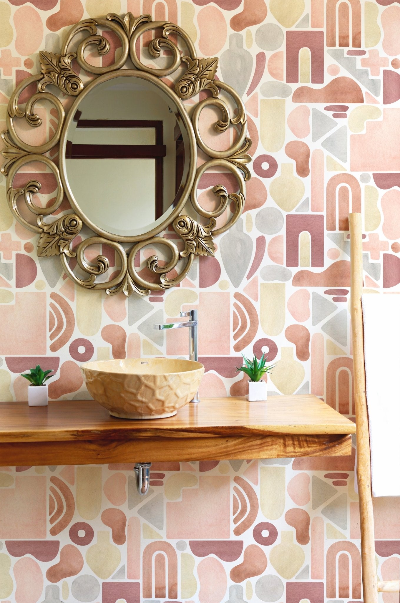 Boho wallpaper in bathroom behind a wooden console table with a gold mirror and small plants.