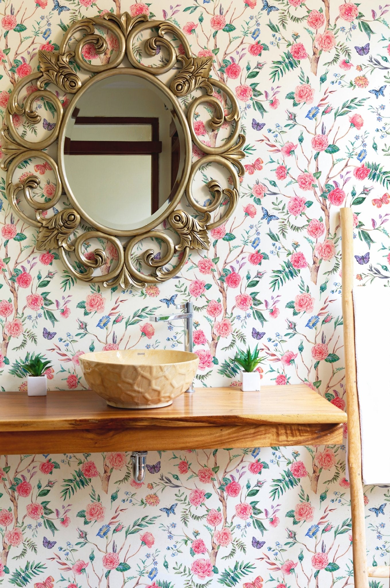 Rose bouquet wallpaper behind a gold ornate mirror on a wooden shelf in bathroom.